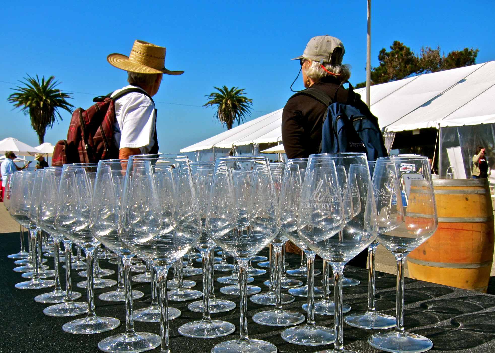 Two people at a wine tasting event with empty wine glasses and tents in the background.