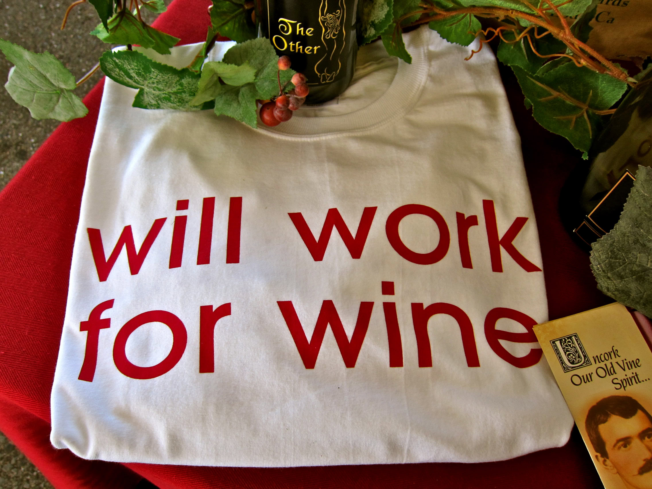 White T-shirt with red text 'will work for wine' on a table with plants and wine-related items.