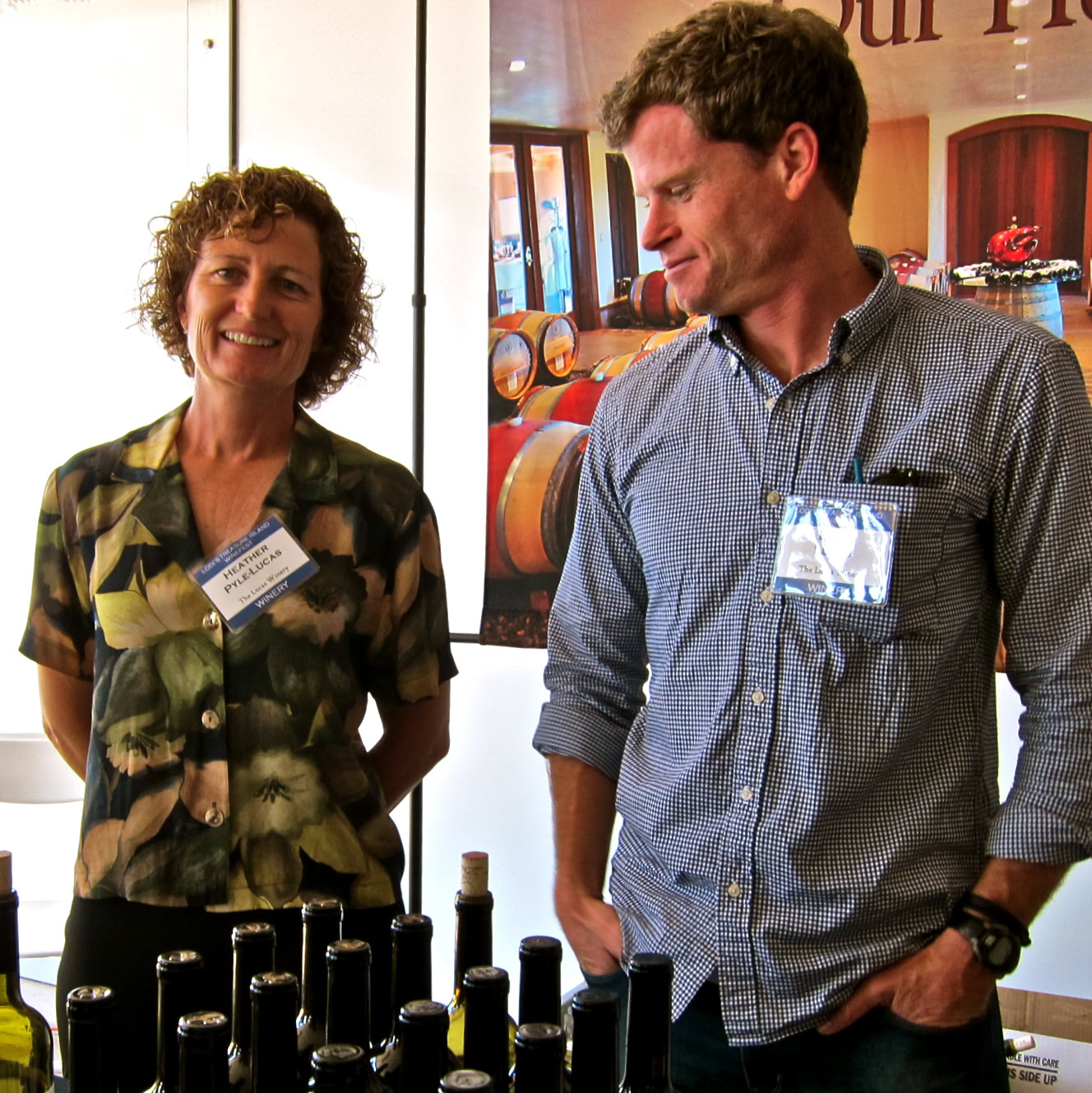 Two people at a wine event with bottles of wine on display, smiling and engaging in conversation.