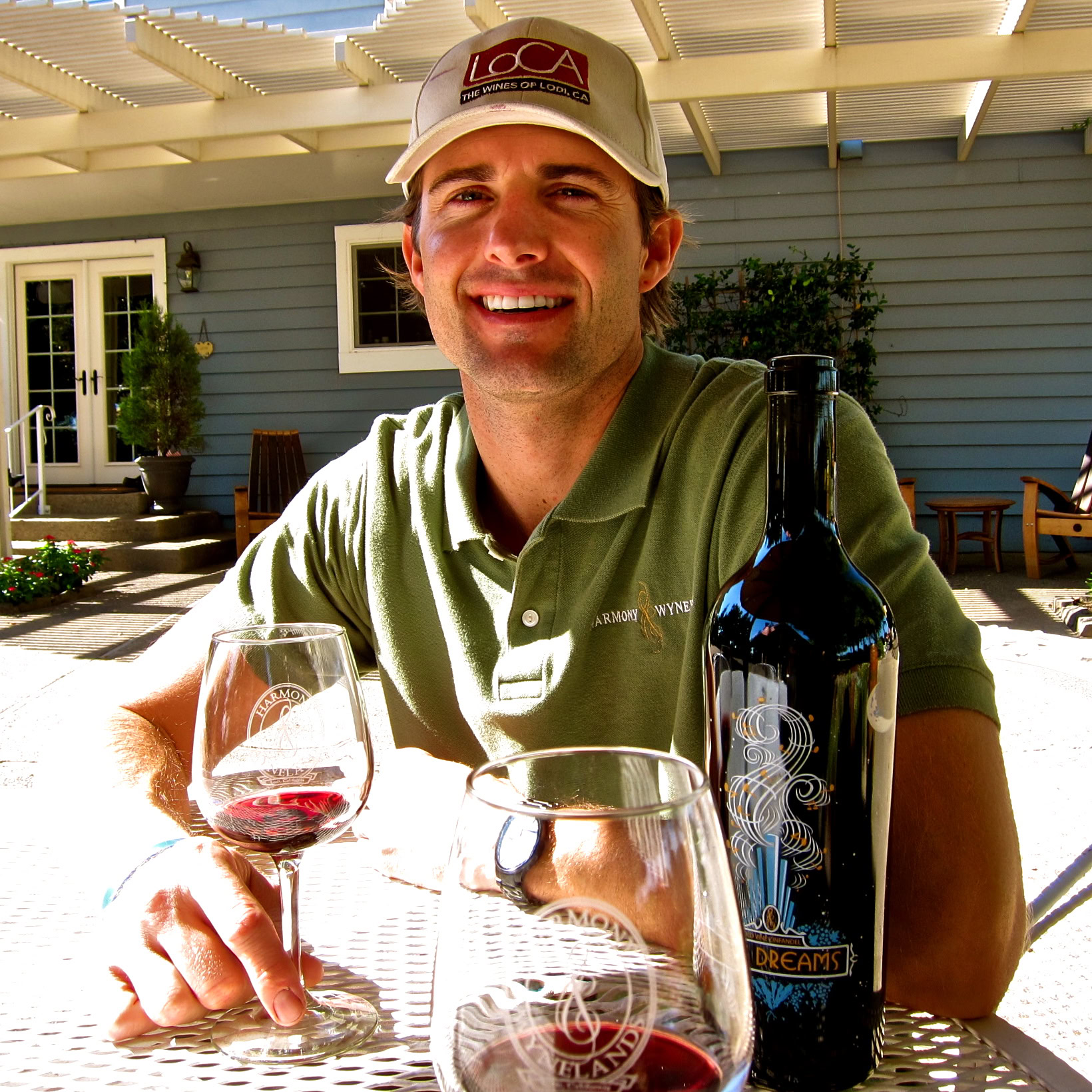 Smiling man in green polo shirt and cap enjoying wine outdoors with wine bottle and glasses.