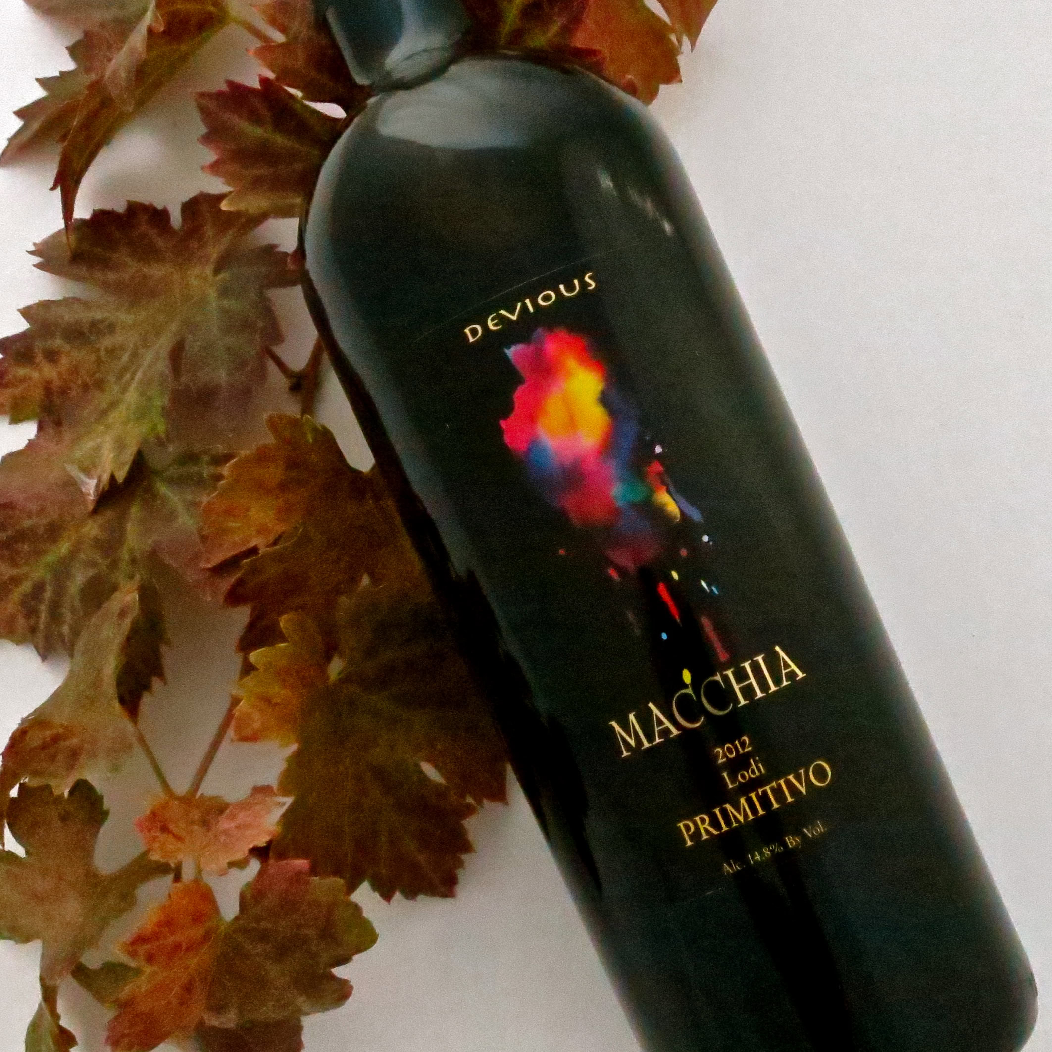 Devious Macchia 2012 Primitivo wine bottle with colorful label, surrounded by autumn leaves.