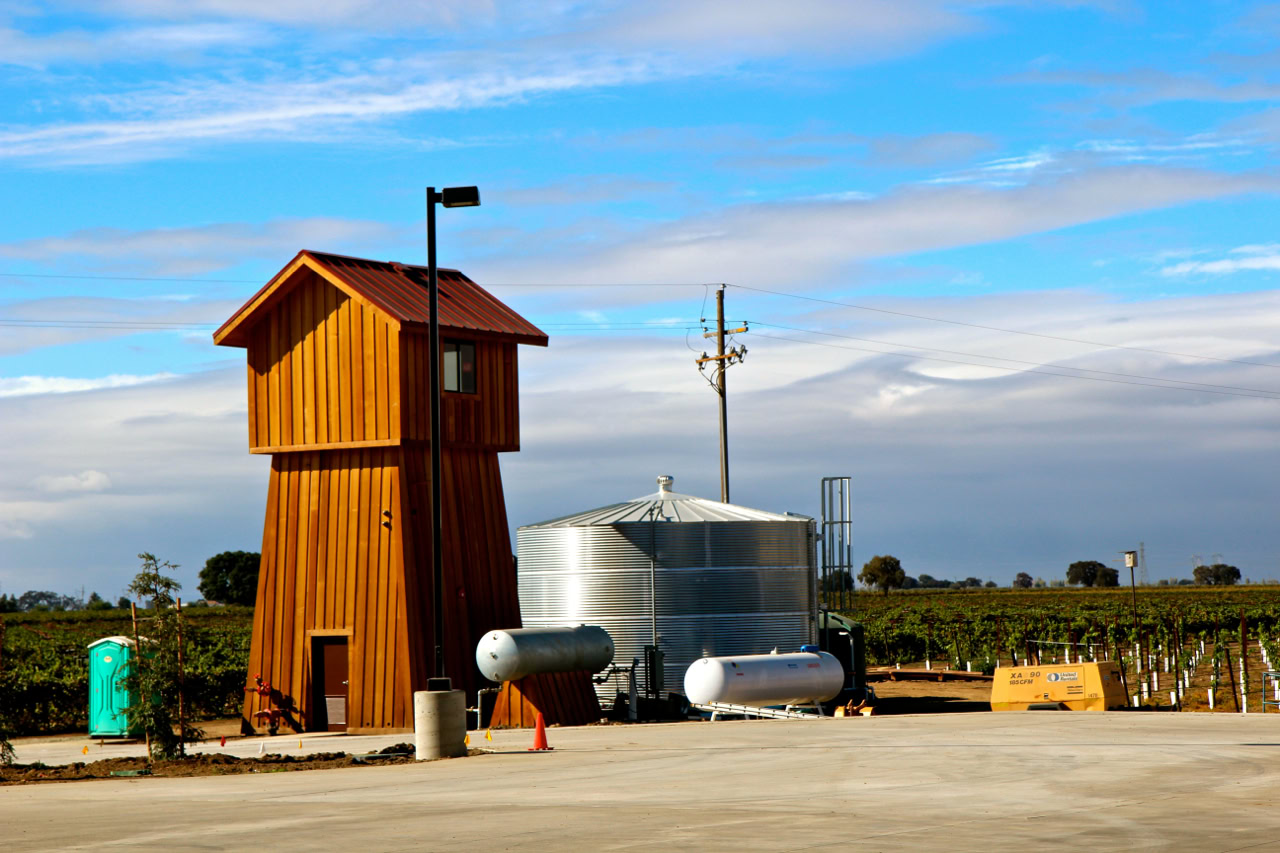 Winegrape irrigation and storage facility in Lodi, California, with a wooden tower and metal tanks under a partly cloudy sky.