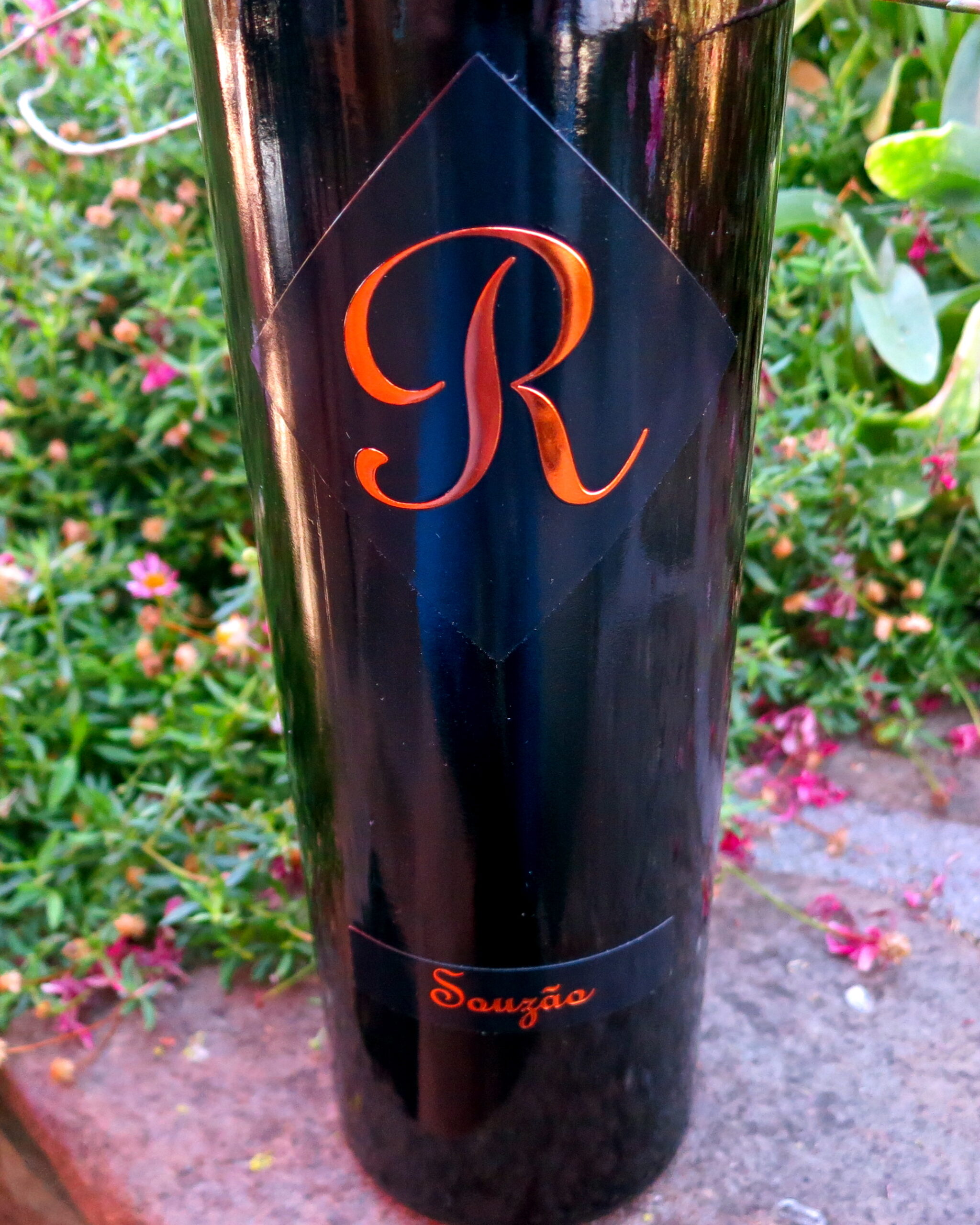 Close-up of a wine bottle with a black label and orange 'R' logo, surrounded by garden plants and flowers.