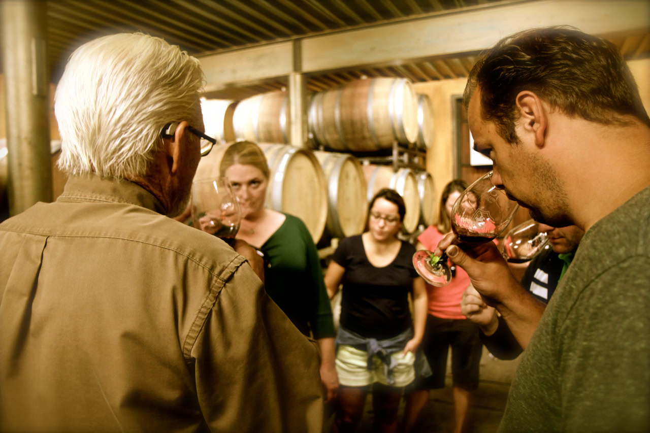 Group of people tasting wine in a winery with wine barrels in the background.