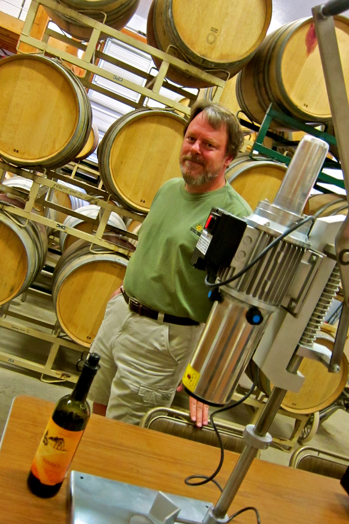 Winemaker in Lodi wine cellar with barrels and wine bottle on table.