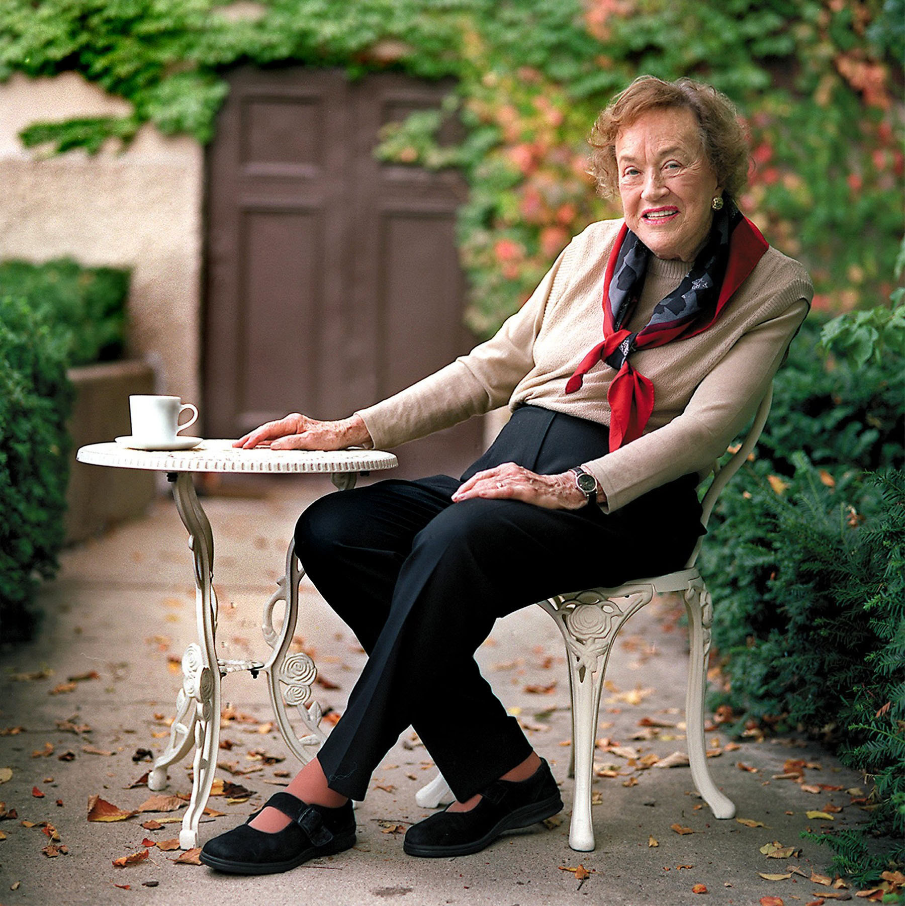 Elderly woman enjoying outdoor tea in a garden with lush greenery and colorful foliage.