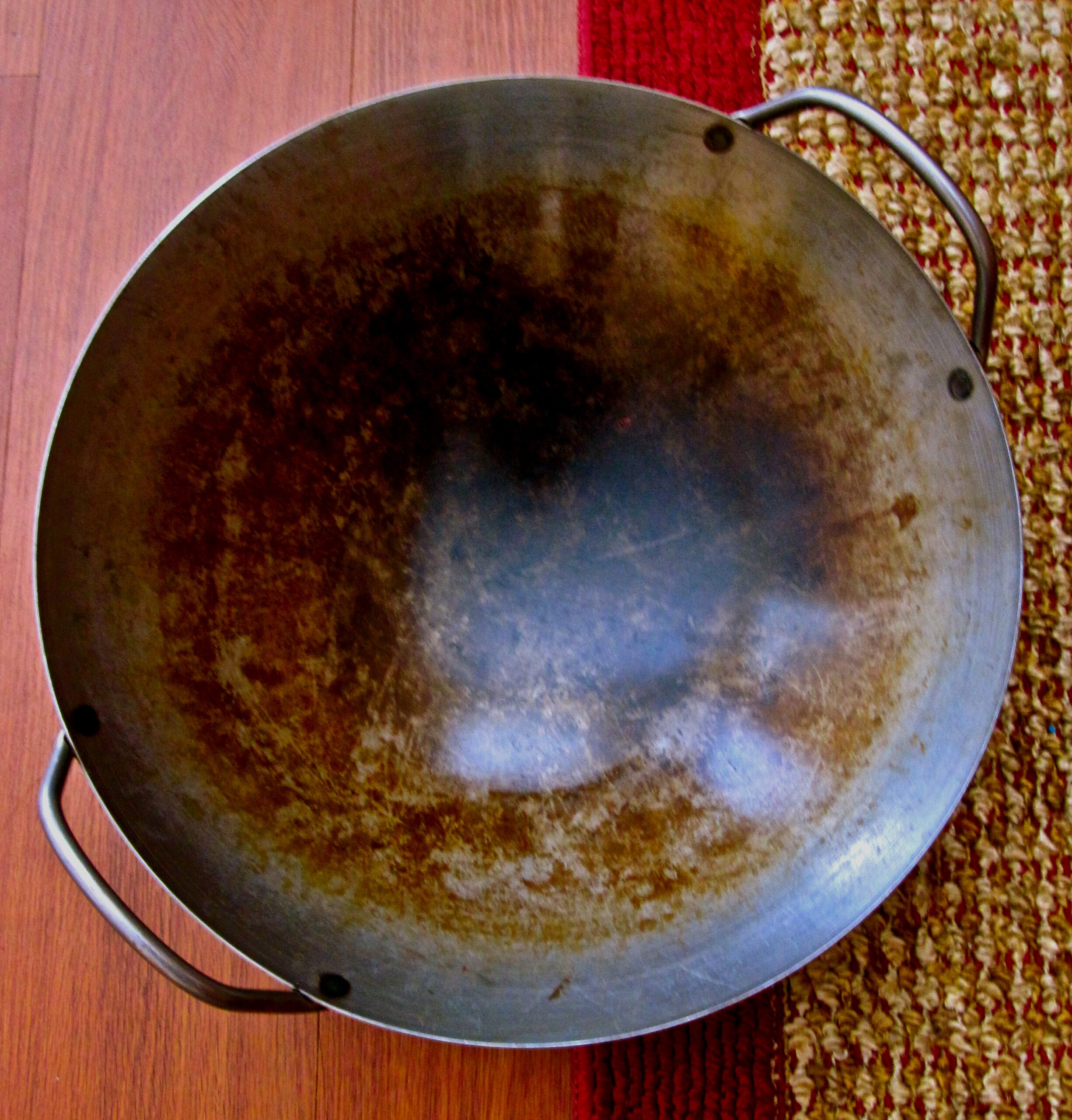Rusty wok with a worn surface, placed on a wooden table with a woven mat nearby.