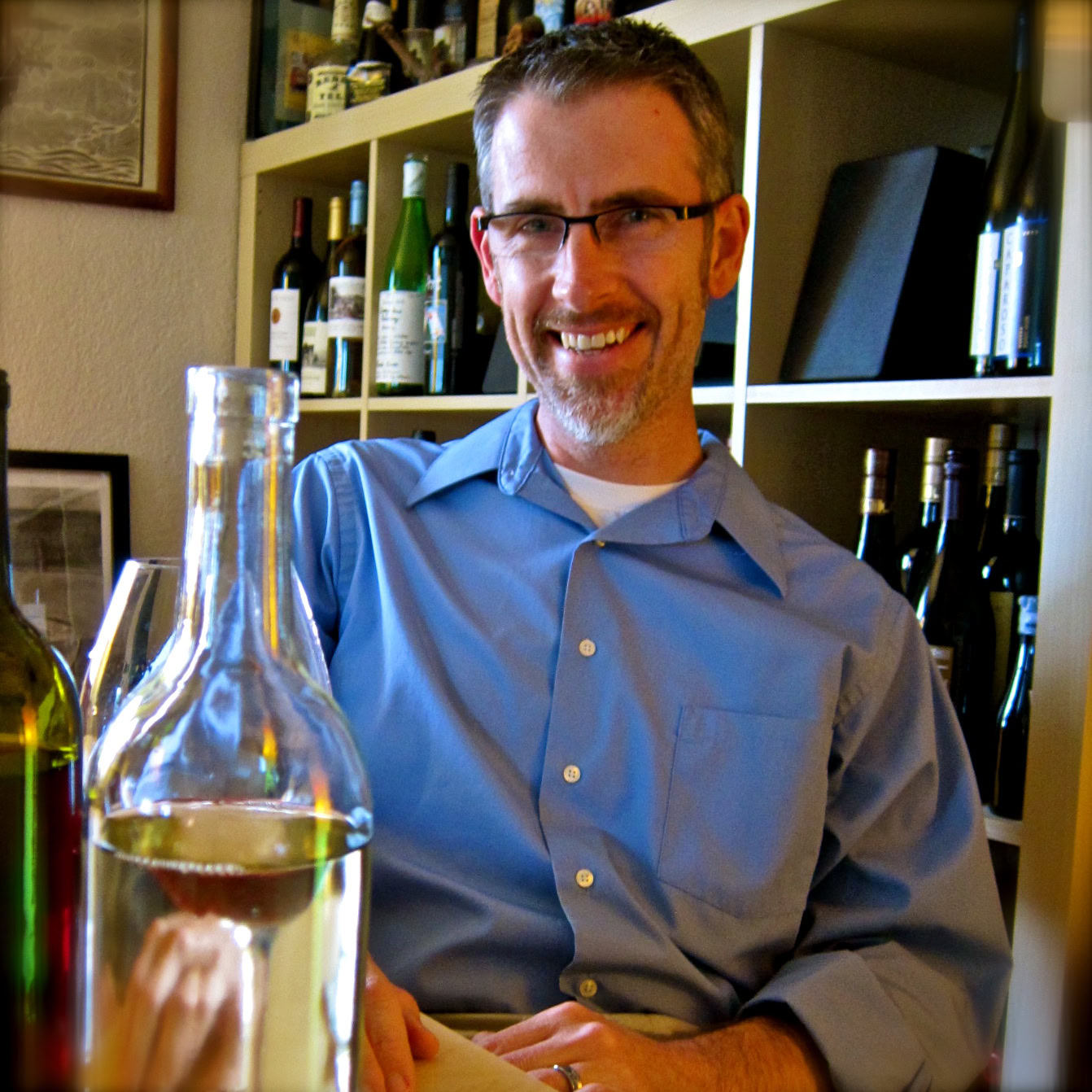Smiling man in blue shirt with wine bottles in background at Lodi Winegrape Commission event.