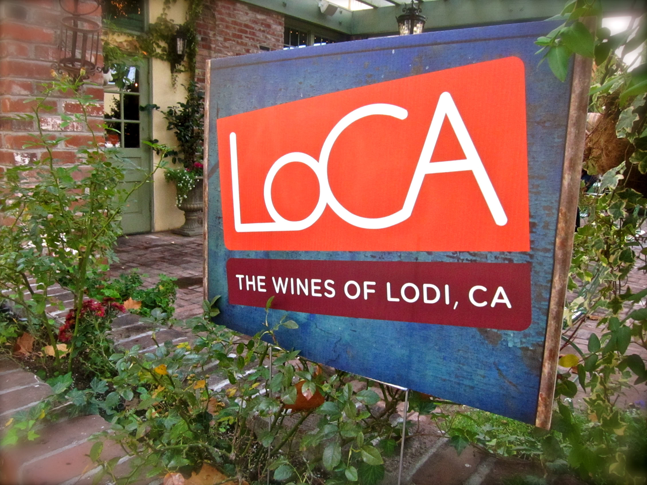 Lodi Winegrape Commission sign in a garden setting with greenery and brick pathway.