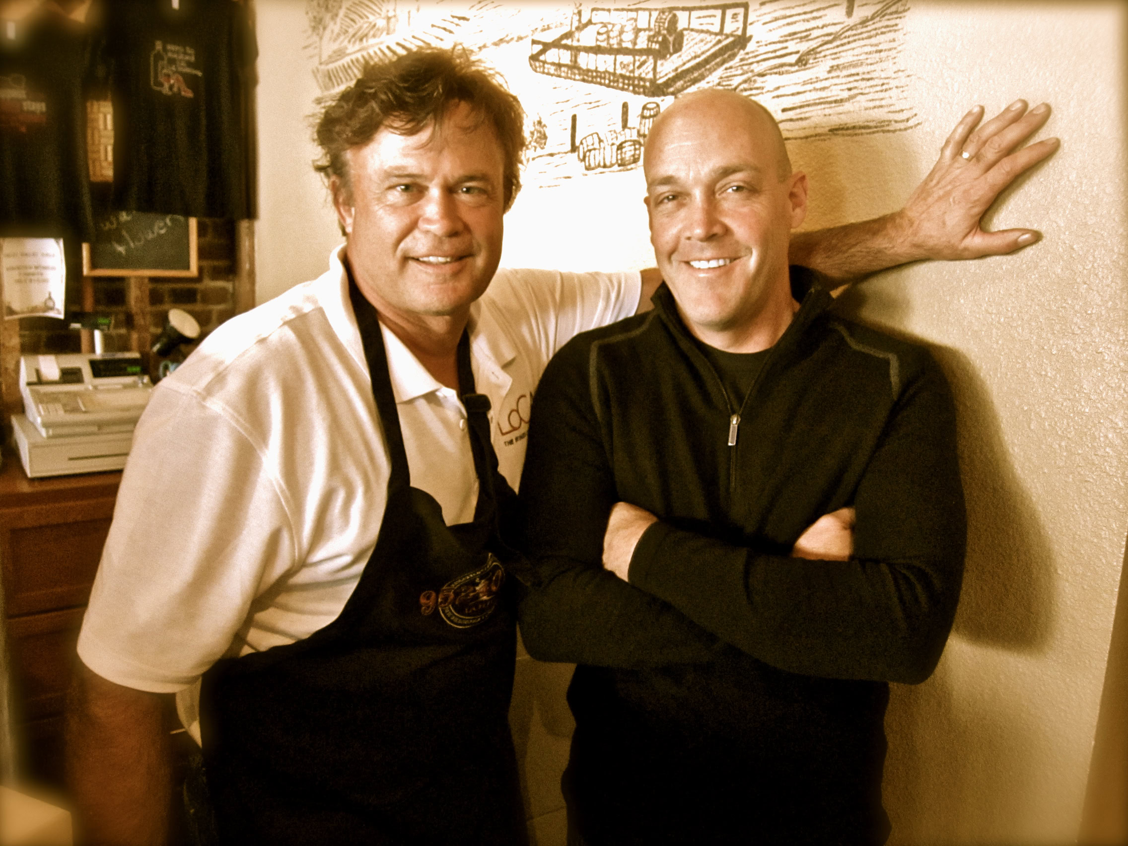 Two men smiling at a Lodi Winegrape Commission event, one wearing an apron, in a cozy wine bar setting.