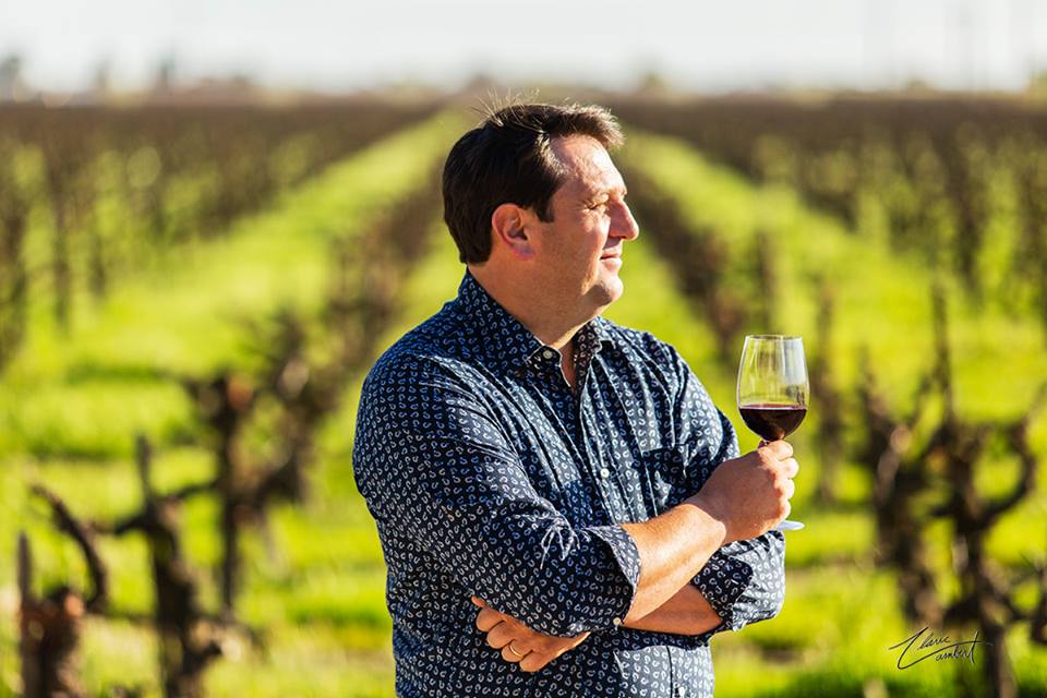 Man holding a glass of red wine in a vineyard with lush green vines in Lodi, California.