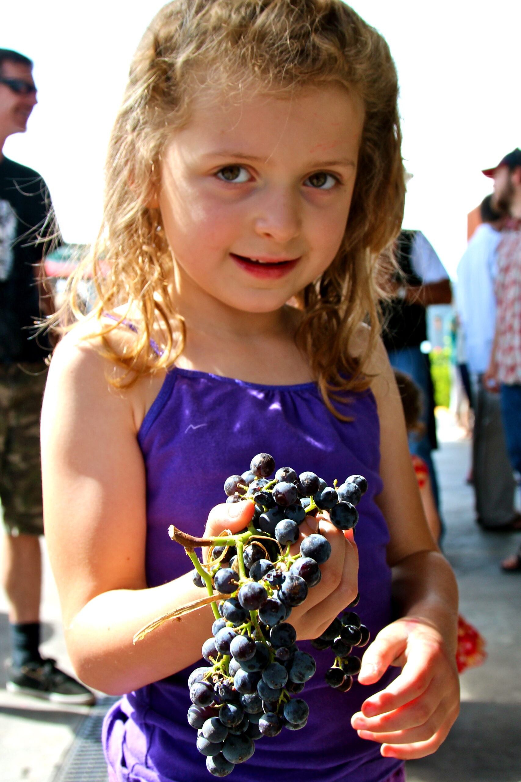 Young girl holding a bunch of ripe grapes at a Lodi wine event, with people in the background.