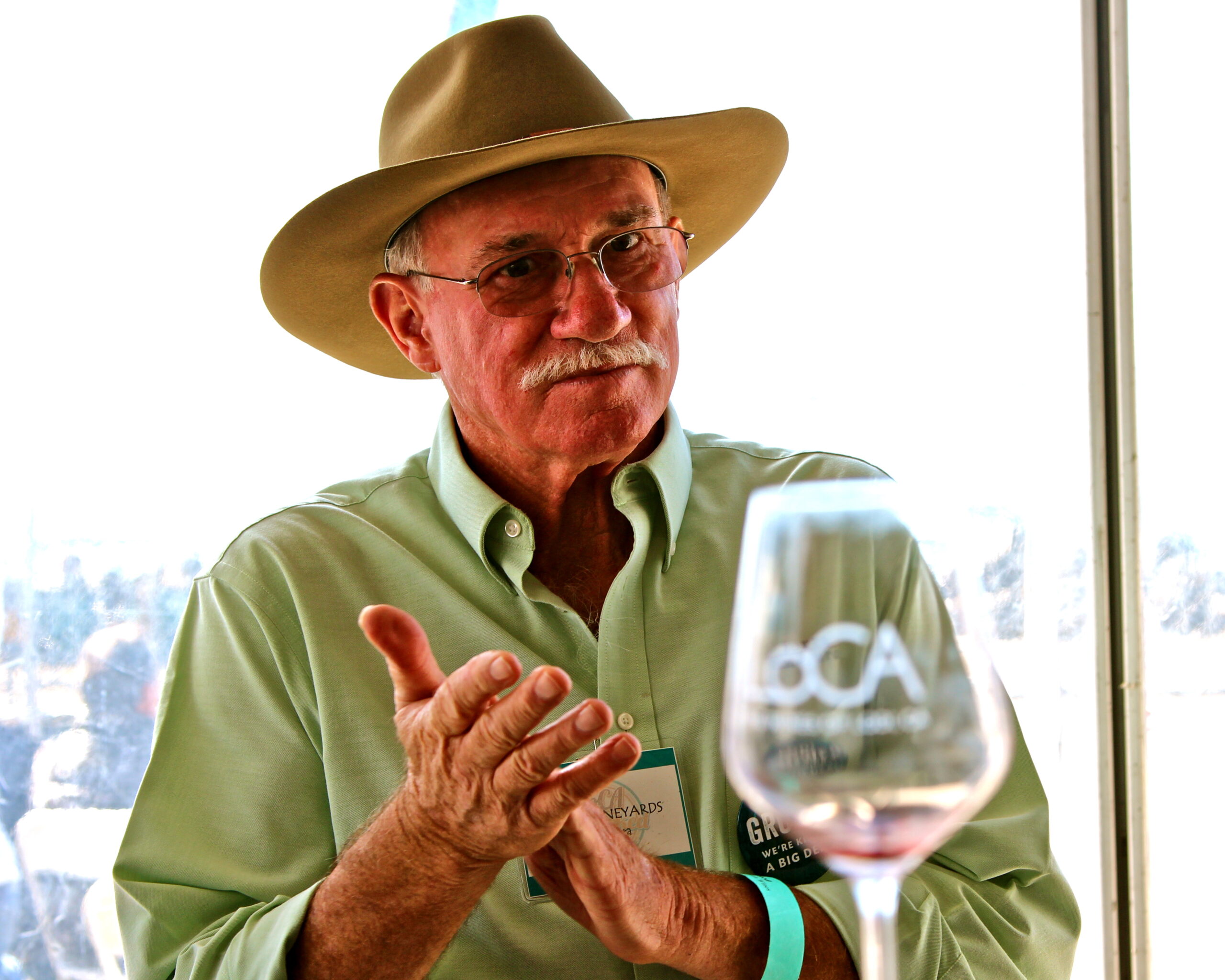 Senior man in hat discussing wine at Lodi Winegrape Commission event with a glass of wine in front.