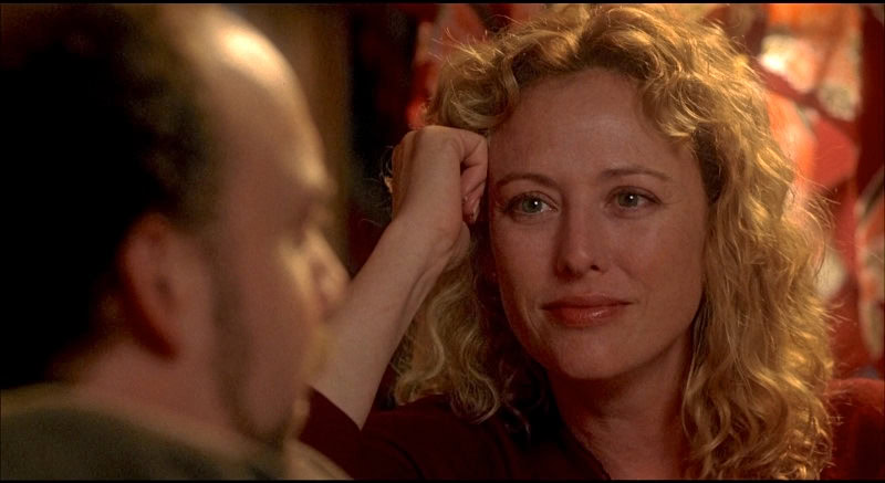 Close-up of a woman with curly blonde hair smiling, with a man in the foreground, in a warm, inviting setting.