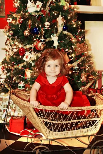 Adorable young girl in red dress sitting in a wicker cradle in front of a decorated Christmas tree.
