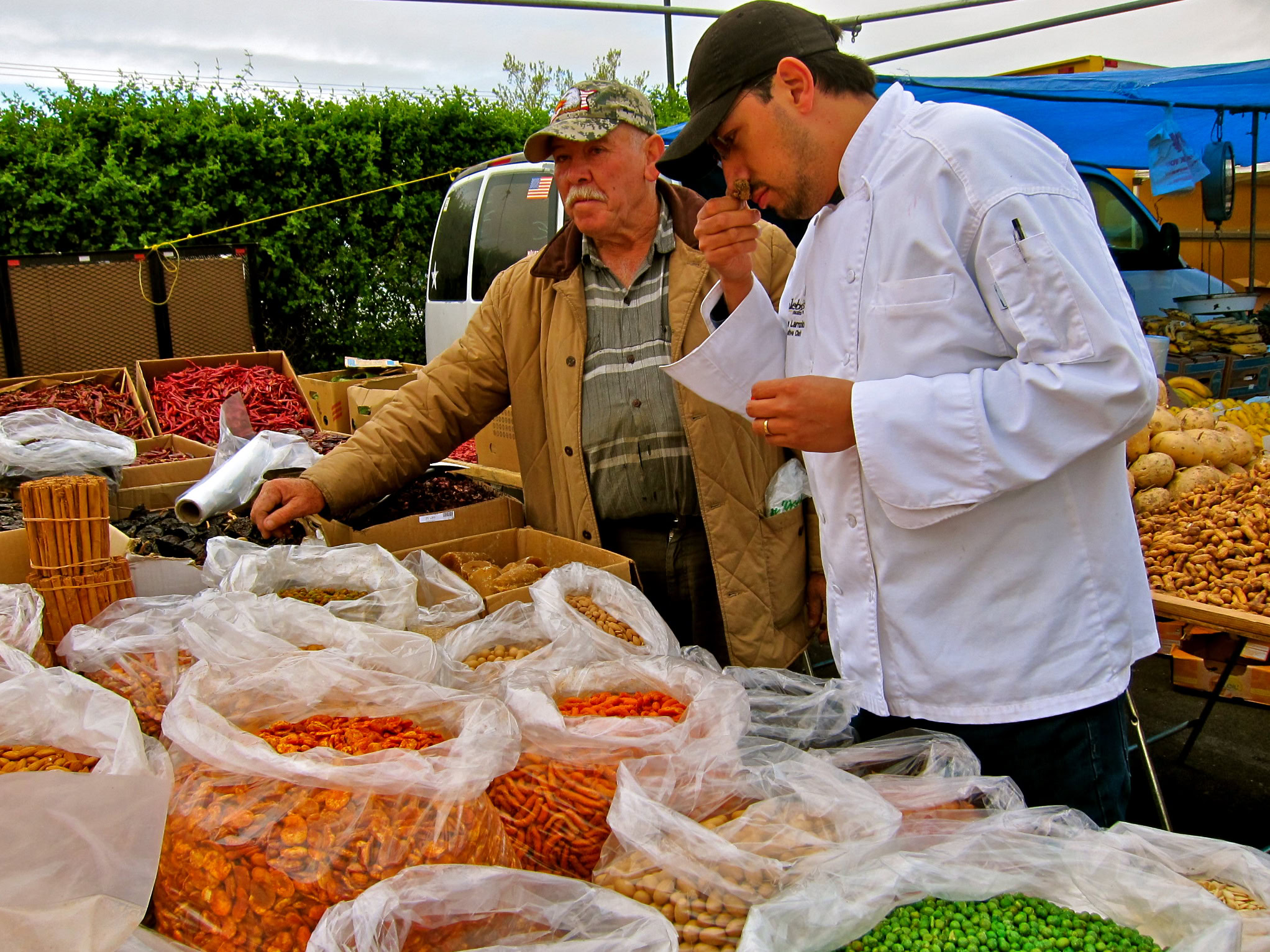 Two men at a market stall with dried fruits and produce, discussing and inspecting items, under a blue canopy.