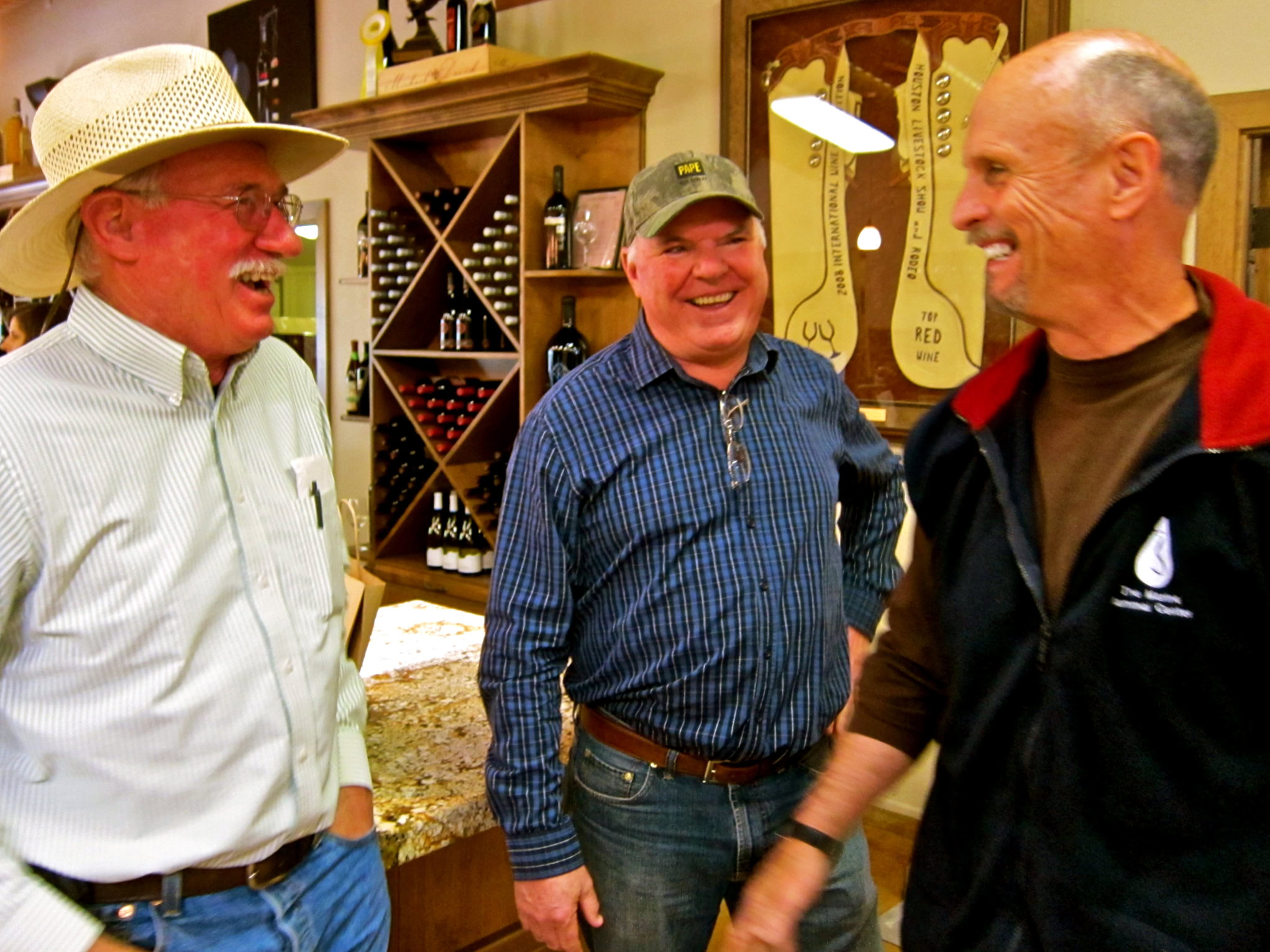Three men smiling and talking in a wine shop, with wine bottles and wine-related decor in the background.