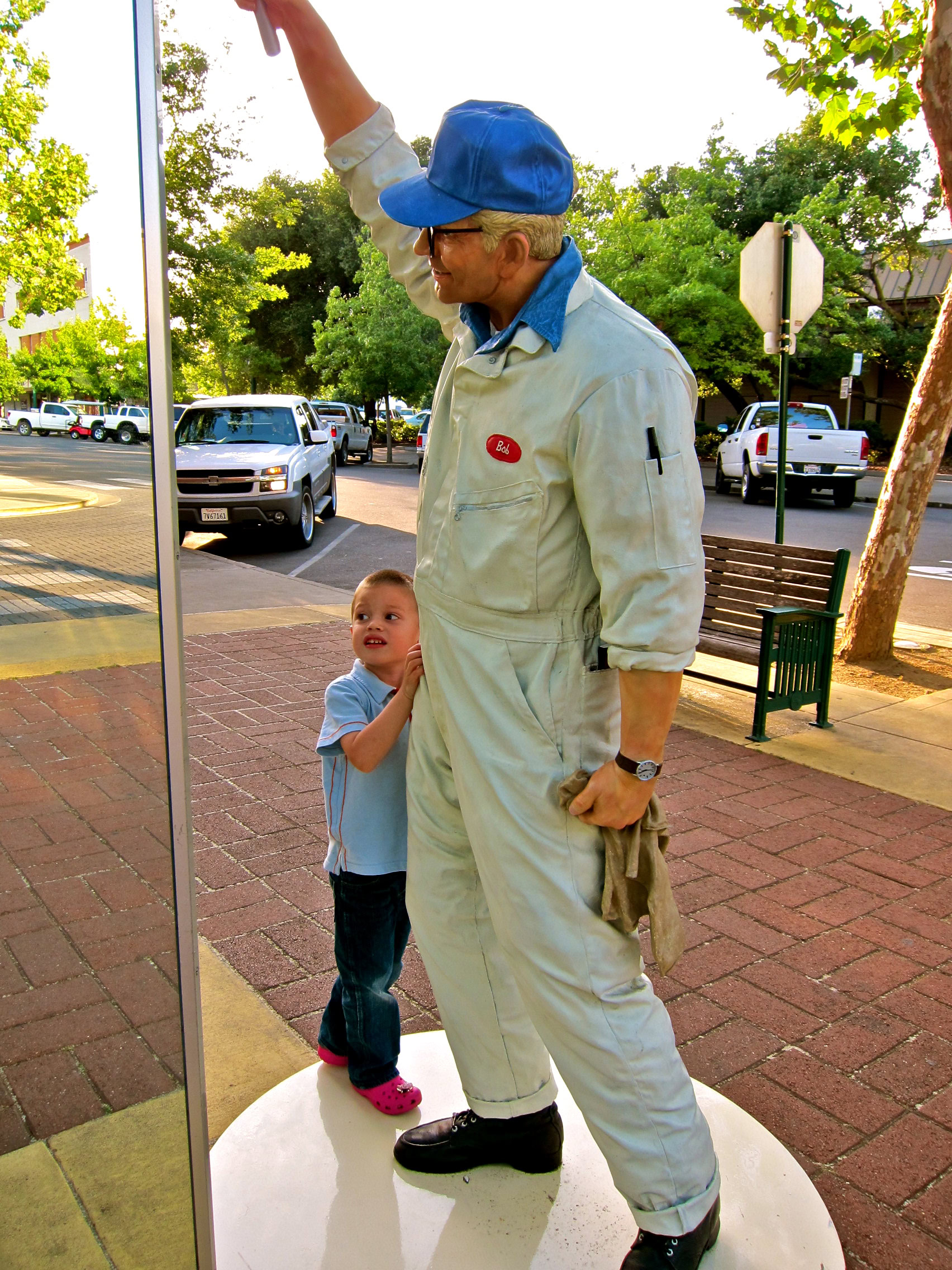 A man in work overalls and a blue cap interacts with a young girl on a city sidewalk.