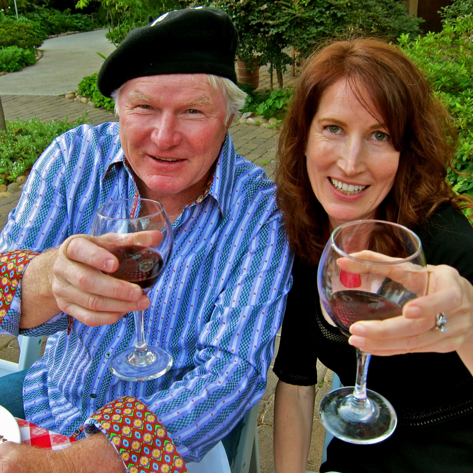 Two people enjoying red wine outdoors, smiling, with lush greenery in the background.