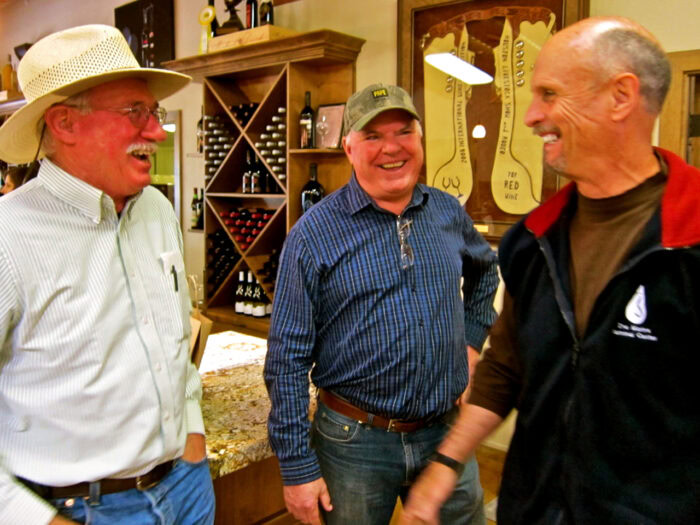 Three men smiling and talking in a wine shop, surrounded by wine bottles and wine-related decor.