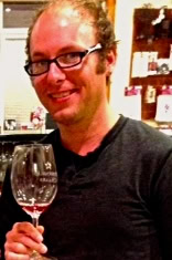 Close-up of a person holding a glass of red wine, smiling, with wine bottles in the background.