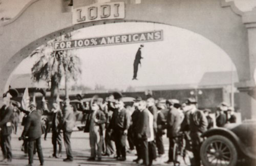Historical black and white photo of people gathered outside a building with a hanging figure and a banner reading 'For 100% A.