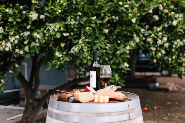 Wine and cheese setup on a barrel with a bottle of wine and a glass in a vineyard setting.