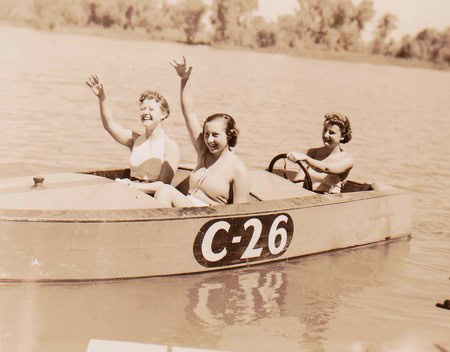 Vintage photo of children in a boat on a lake, smiling and waving, sepia tone, nostalgic scene.