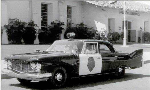 Black and white vintage police car with siren and shield emblem parked on street in Lodi.