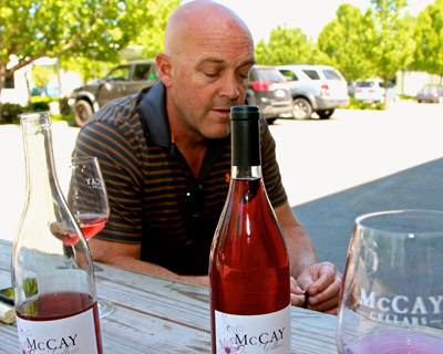 Man tasting wine outdoors with bottles and glasses on table, lush green trees in background.