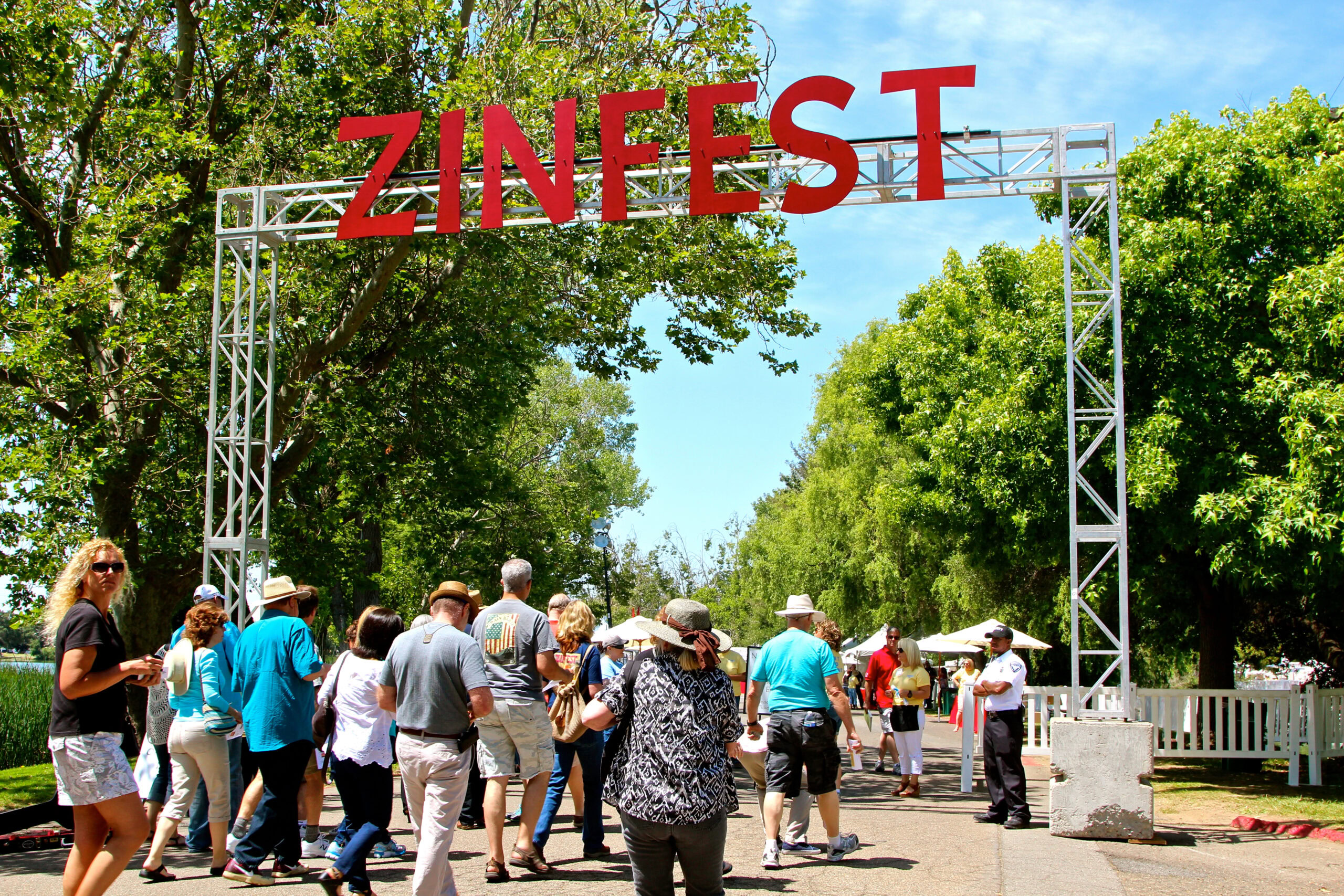 Visitors at Zinfest event under a large sign with trees and blue sky in the background.