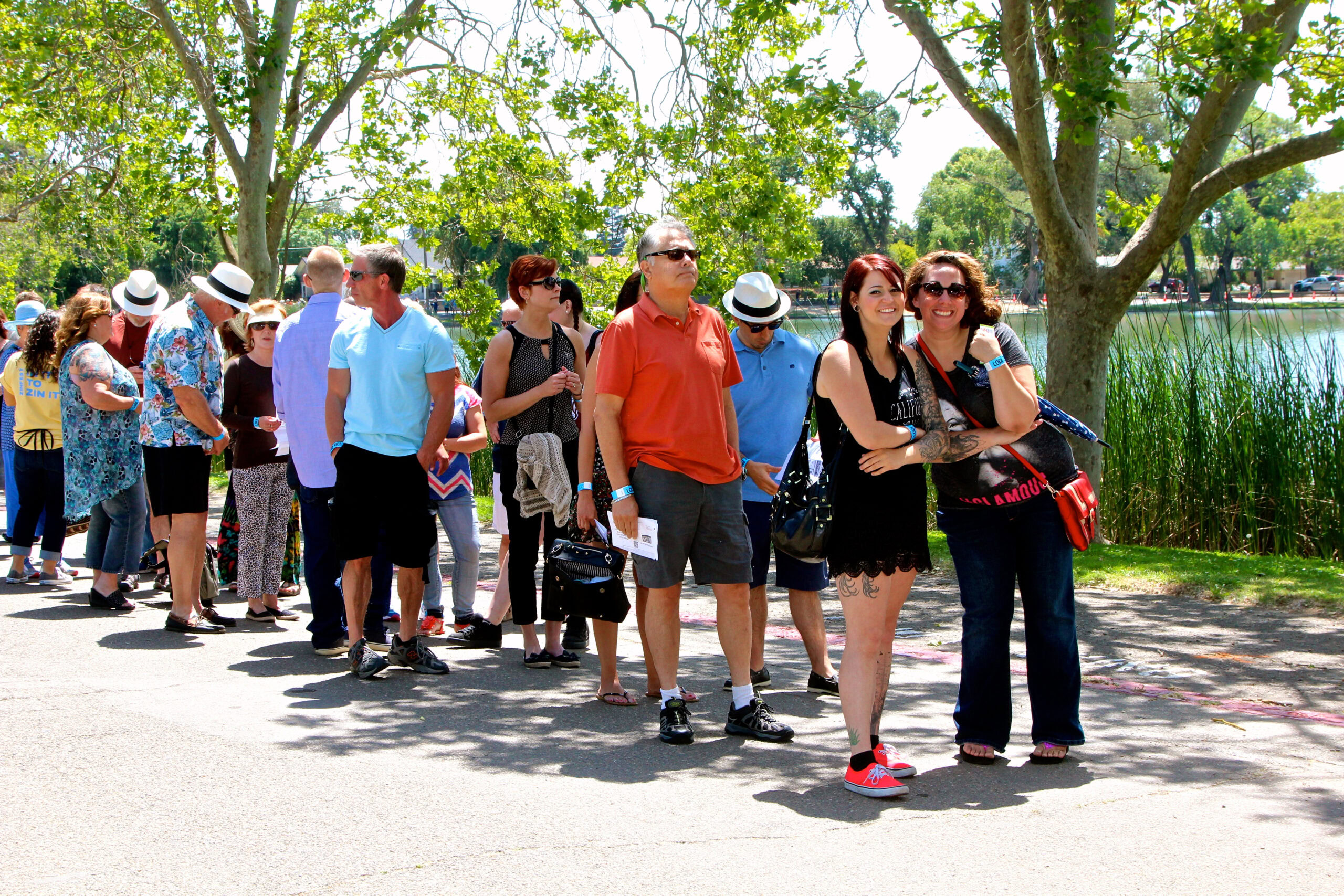 Tour group waiting in line outdoors near trees and water at Lodi Winegrape Commission event.