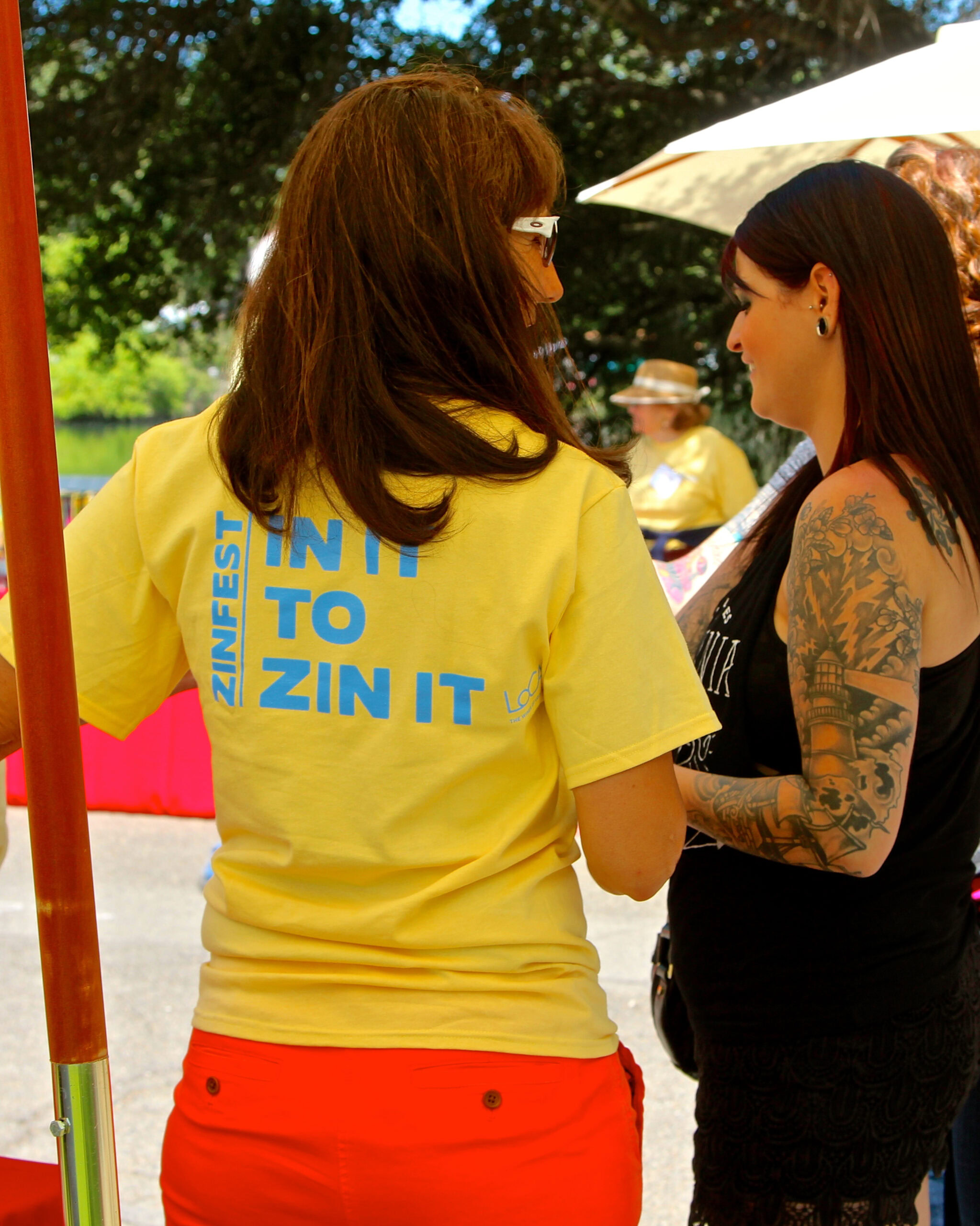 Two women talking at a Lodi Winegrape Commission event outdoors with trees and umbrellas in the background.