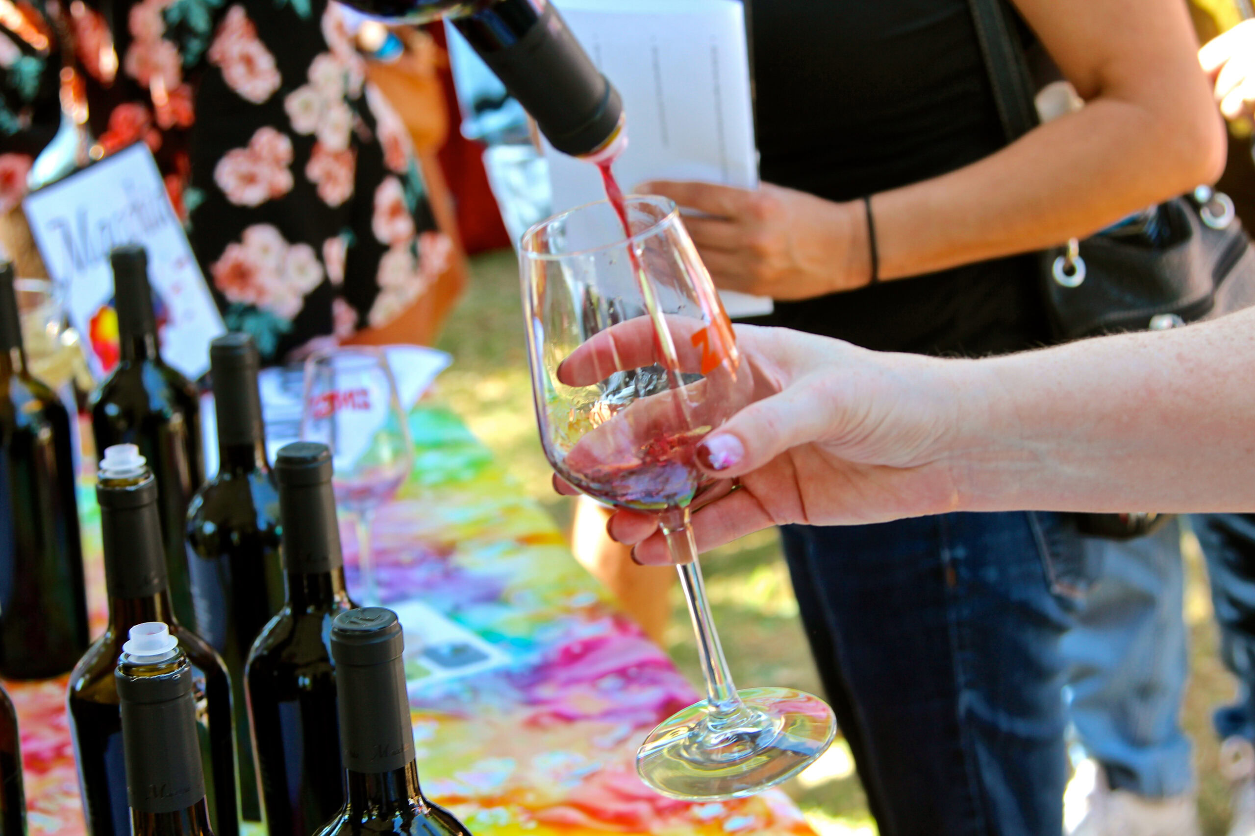 Person pouring red wine into a glass at a wine tasting event with bottles on a colorful table.