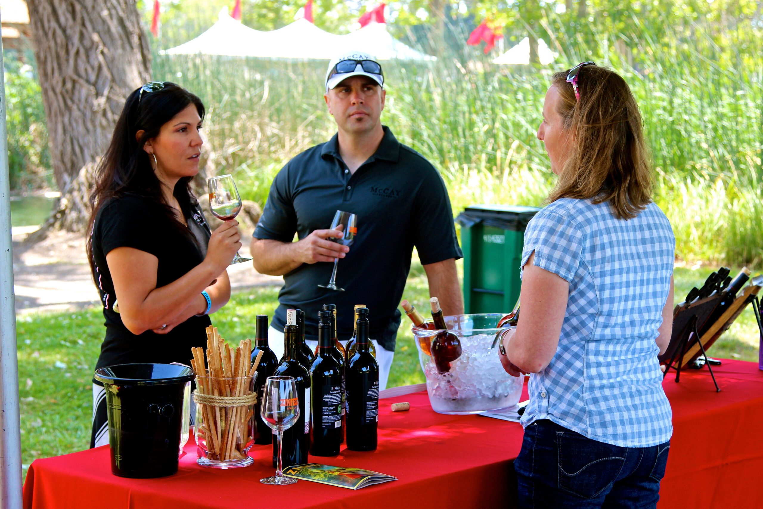 People enjoying wine tasting at Lodi Winegrape Commission event outdoors with wine bottles and glasses on a red table.