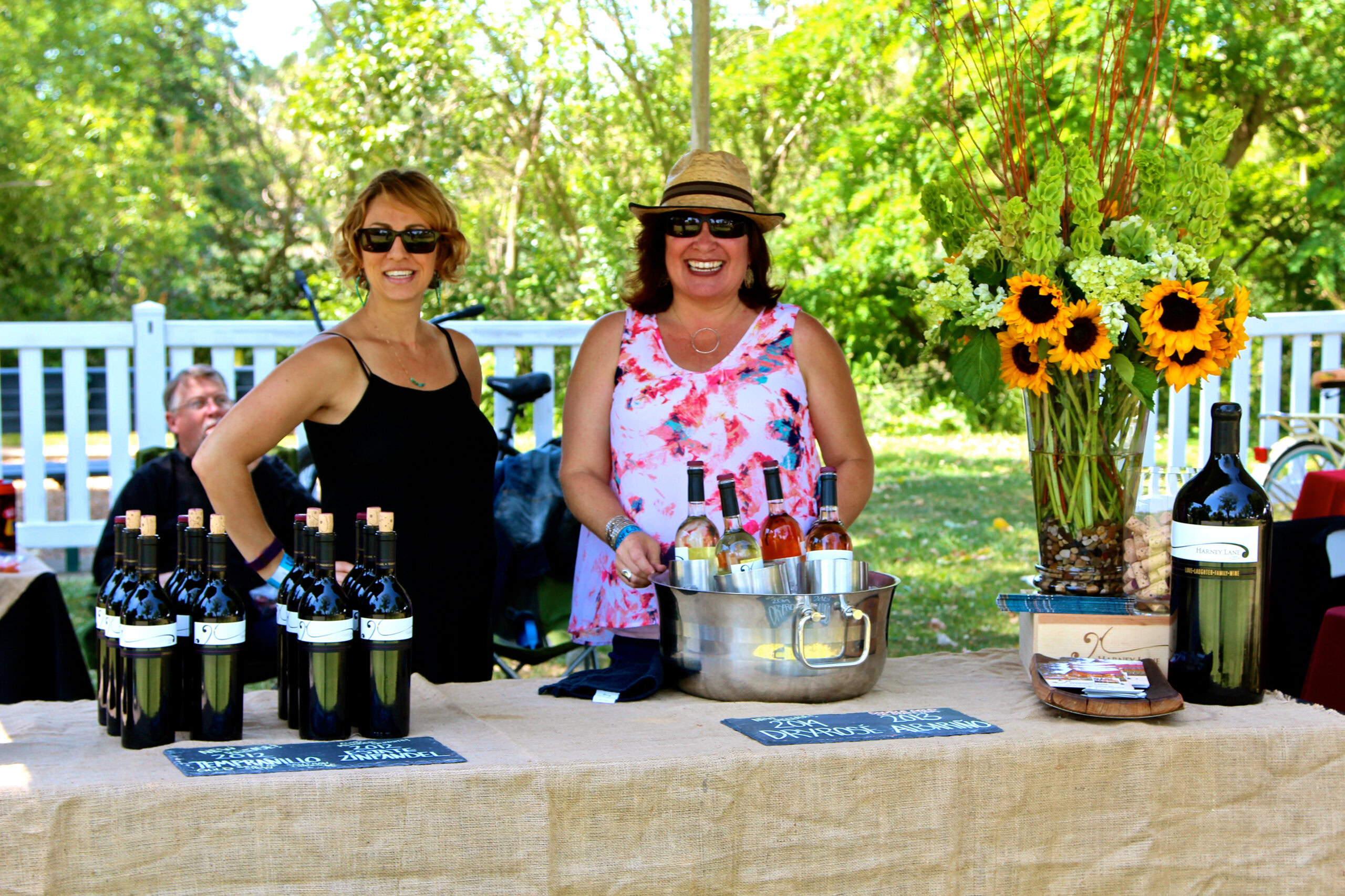 Two women at a wine tasting booth with bottles of wine and sunflowers, outdoors in a lush green setting.