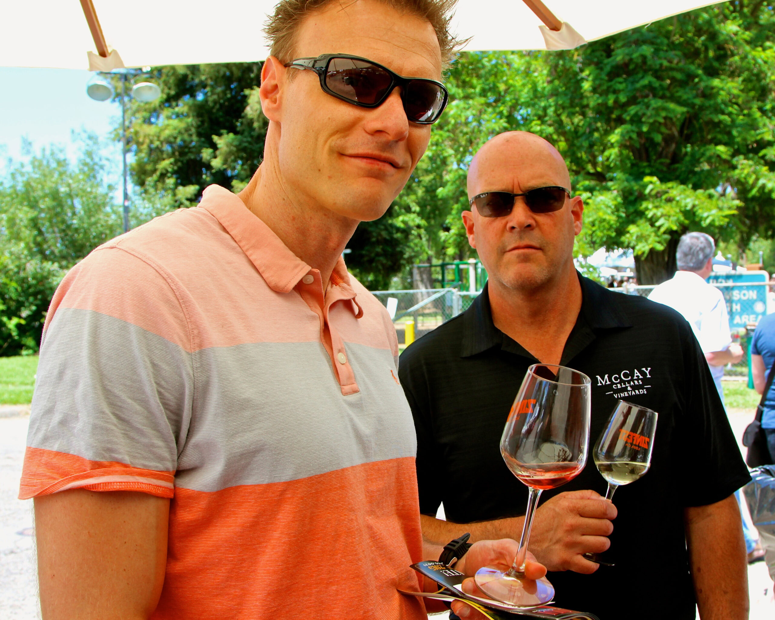 Two men at a wine tasting event holding glasses of red wine outdoors with green trees in the background.