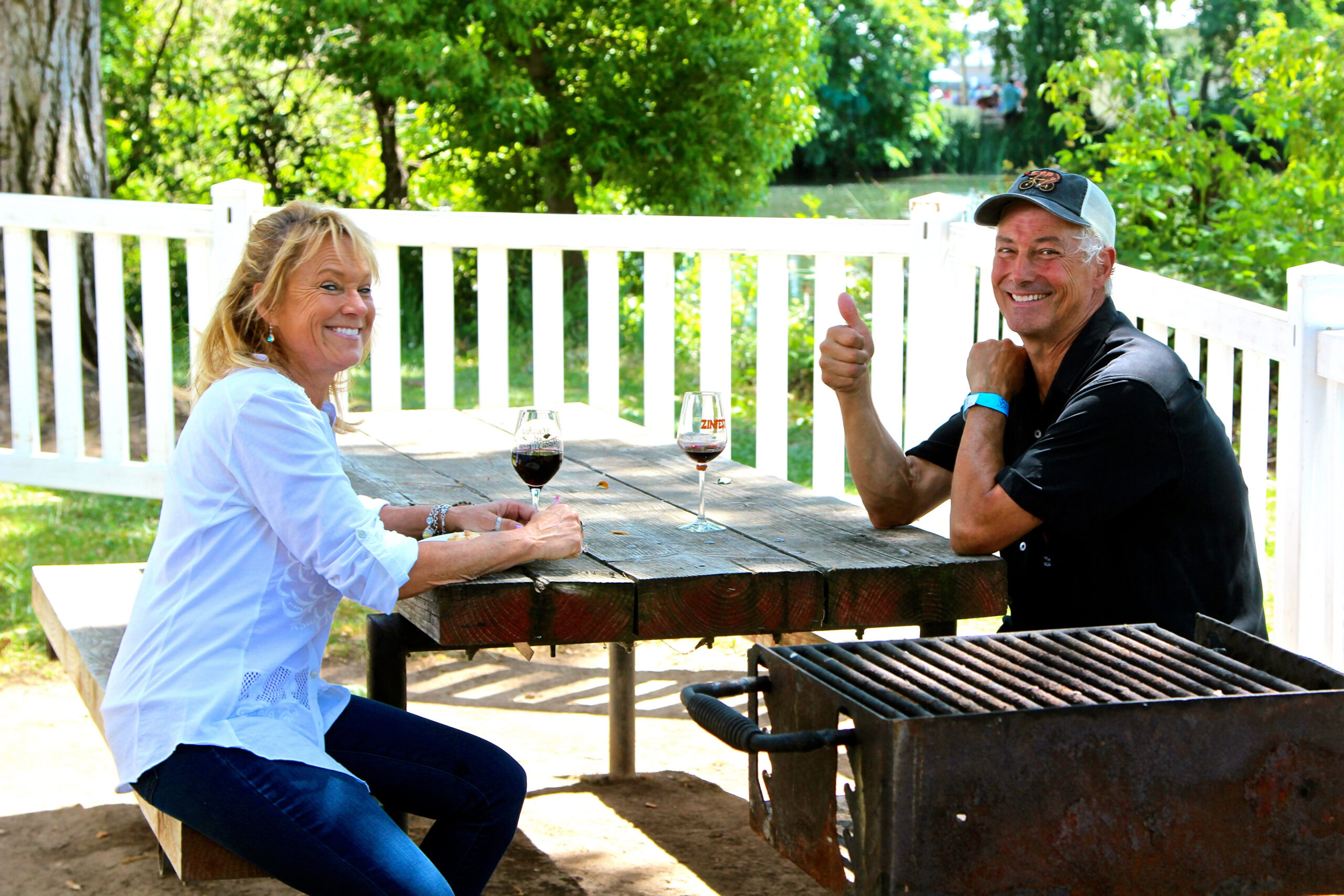 Two people enjoying wine tasting outdoors at a Lodi vineyard with a white fence and lush greenery.
