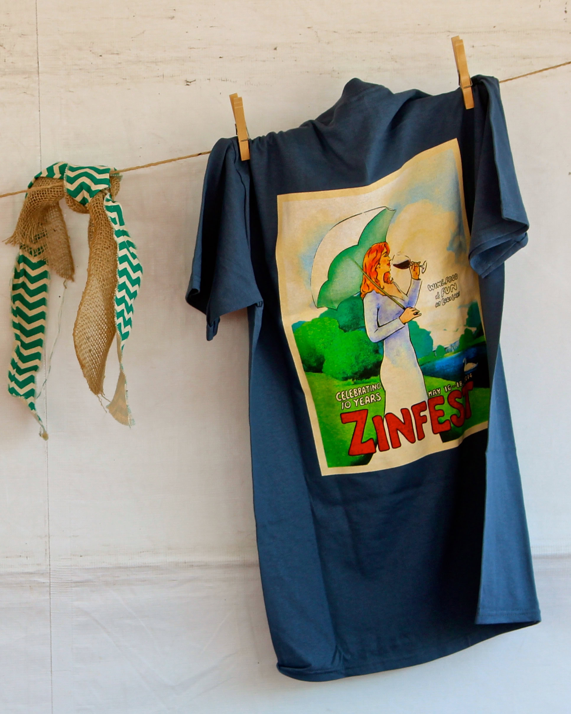 Blue T-shirt with Zinfandel celebration graphic hanging on a clothesline with decorative fabric strips.