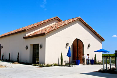 Lodi Winegrape Commission winery with white walls, red tile roof, and outdoor seating area under blue sky.