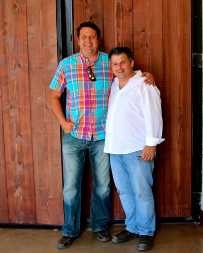 Two men smiling and posing together at a Lodi Winegrape Commission event, standing in front of a wooden wall.