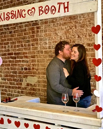 Couple kissing at a romantic kissing booth with heart decorations, brick wall background, and wine glasses on the table.