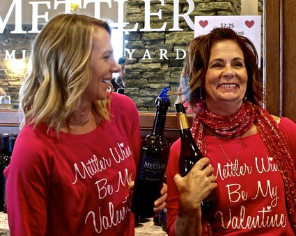 Two women in red shirts celebrating Valentine's Day with wine bottles at Lodi Winegrape Commission event.