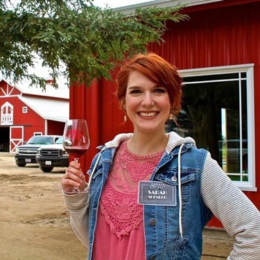 Smiling woman holding a glass of red wine at a Lodi wine event with a red barn in the background.