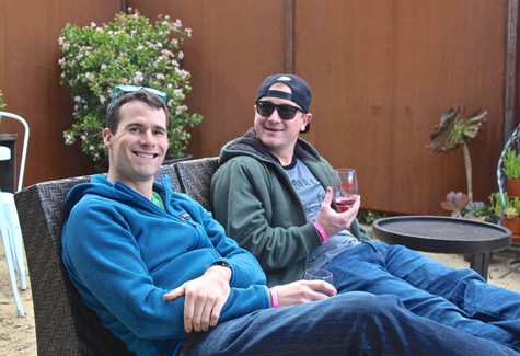 Two men relaxing outdoors with wine glasses, surrounded by plants and wooden fencing.