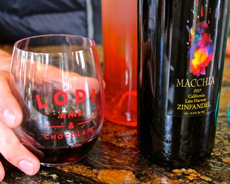 Glass of red wine with Lodi Winegrape Commission logo and wine bottles on a bar counter.