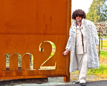 Person in stylish coat and wig standing next to Lodi Winegrape Commission sign.