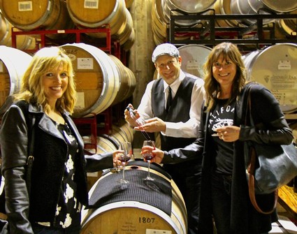 Group of three people enjoying wine tasting at a winery with wine barrels in the background.