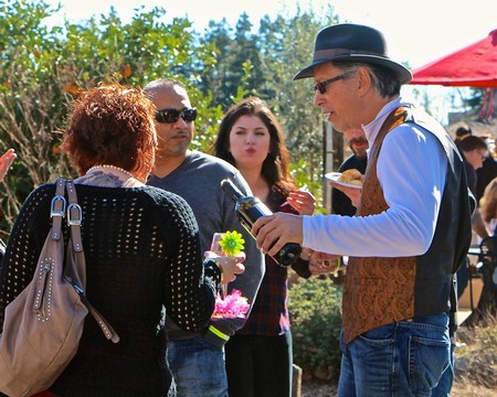 Man in hat and vest speaking to a group at a Lodi wine event, outdoors with trees and a red umbrella in background.