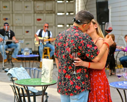 Romantic couple sharing a kiss with wine glasses at outdoor wine tasting event.
